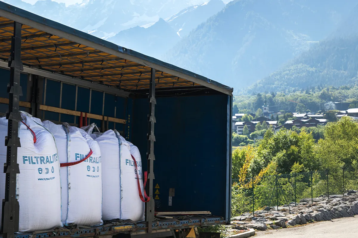 Grands sacs Filtralite chargés dans un camion ouvert, avec un village de montagne et des collines boisées en arrière-plan