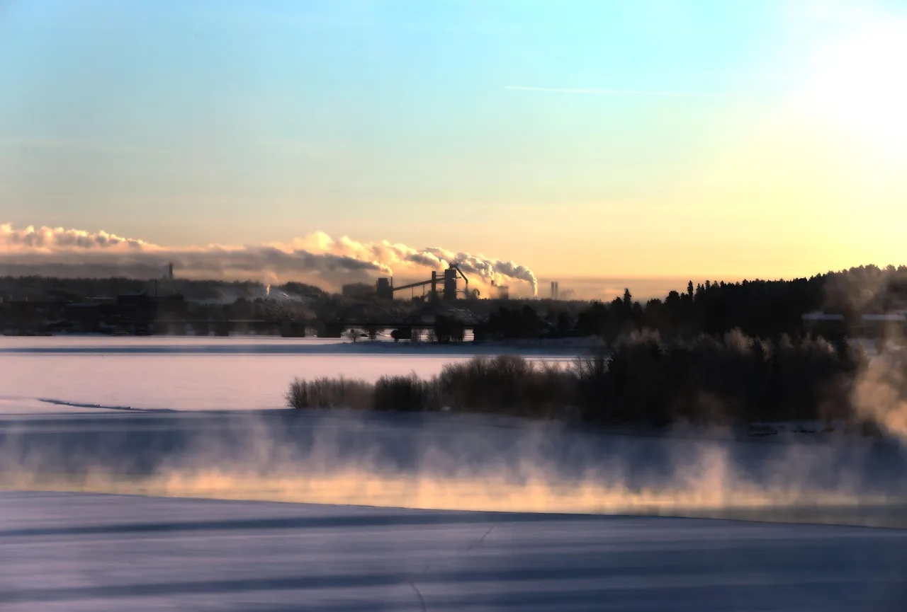 Zone industrielle au loin à Luleå, en Suède, au lever du soleil, avec du brouillard s’élevant au-dessusd’une rivière gelée