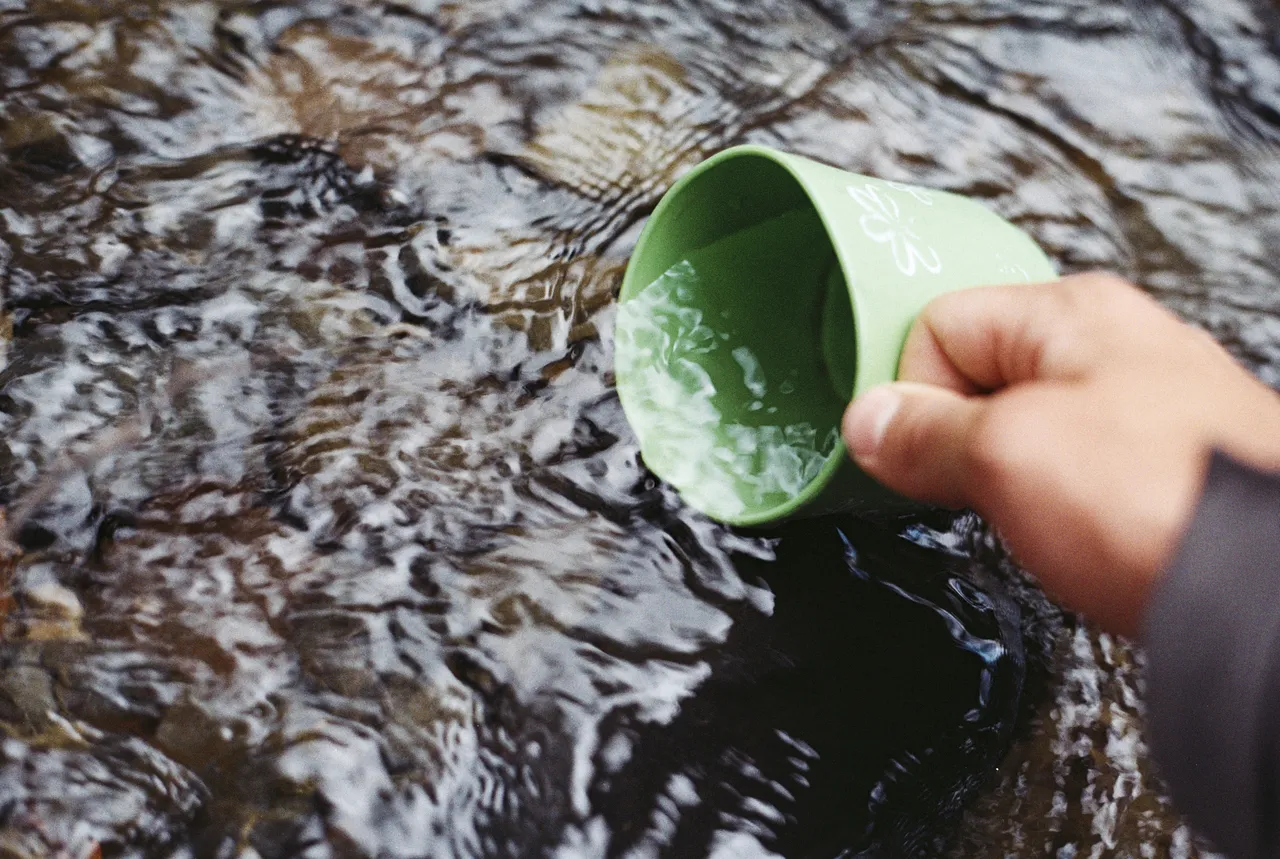 Gros plan d’une personne recueillant de l’eau d’un ruisseau à l’aide d’un gobelet vert.